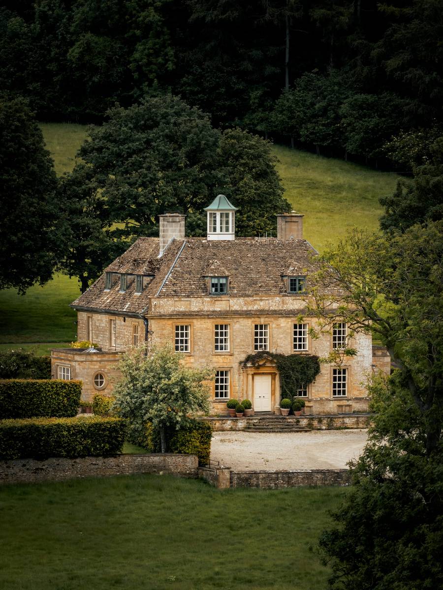 Honey-stone Cotswold manor house framed by clipped hedges and woodland